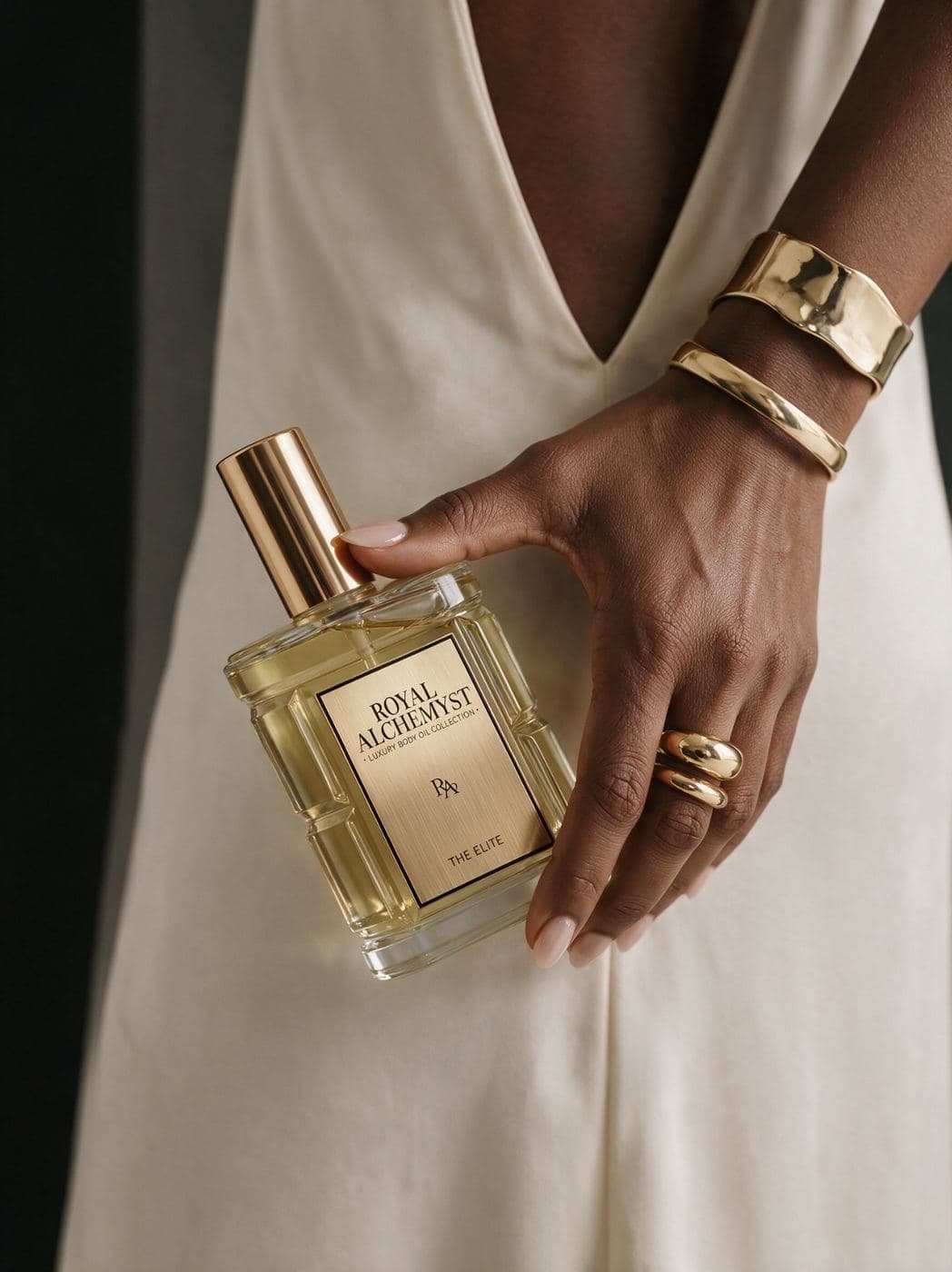 A hand holding The Elite body oil against a cream silk dress